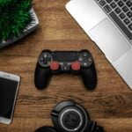 itens de entretenimento, um controle de videogame, fones de ouvido, e eletrônicos sobre uma mesa de madeira.